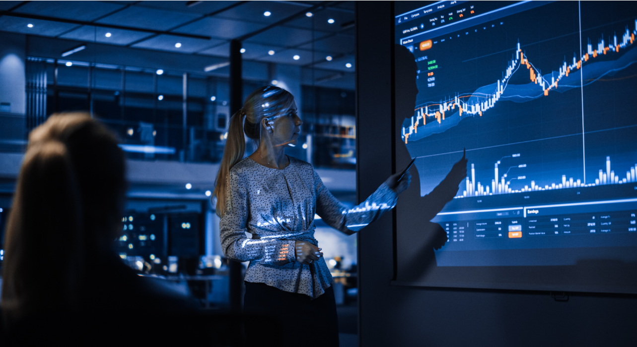 Uma mulher num escritório gesticula em direção a um grande ecrã que exibe gráficos financeiros dinâmicos